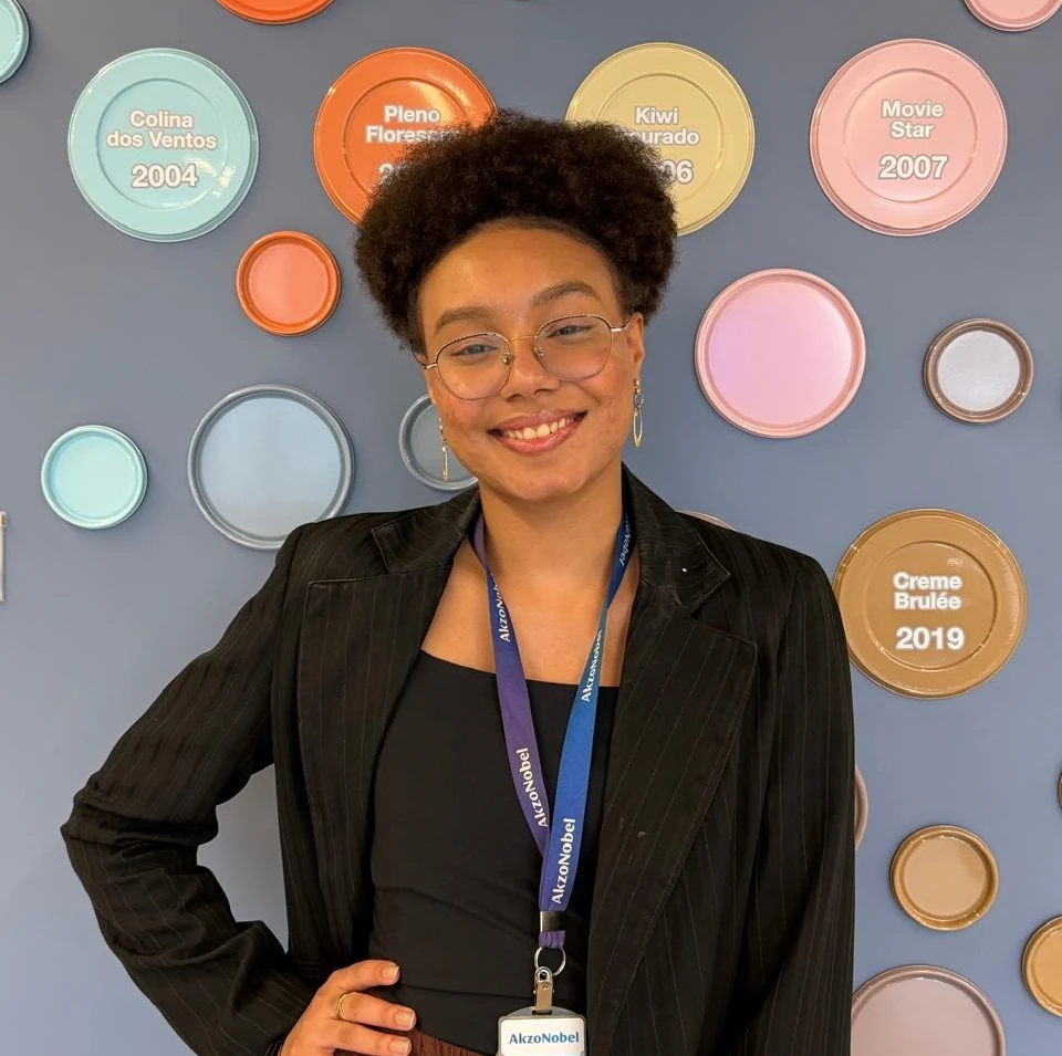 Paula Oliveira sorrindo diante de uma parede com latas de tinta coloridas