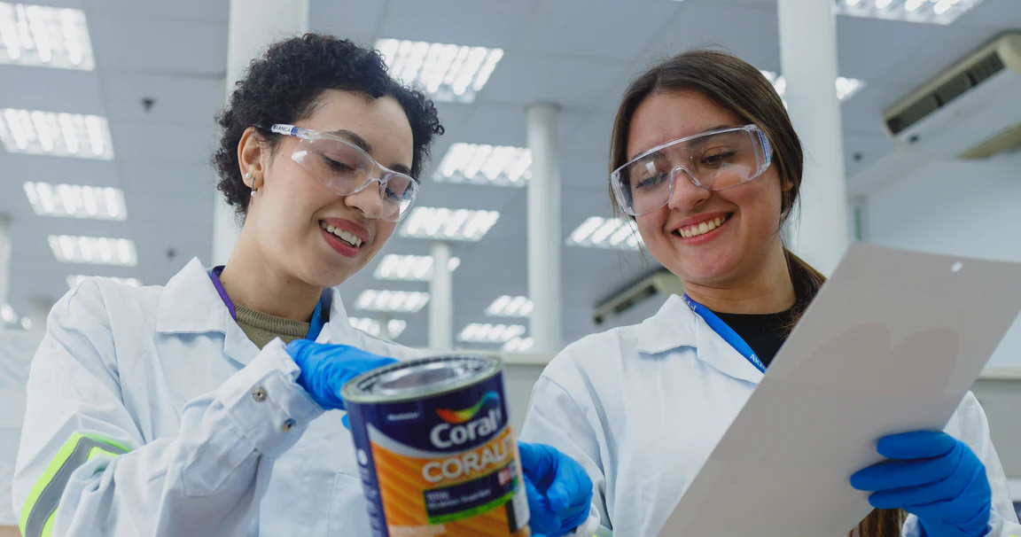 Duas pesquisadoras de jaleco e óculos de proteção sorrindo no laboratório da AkzoNobel enquanto analisam uma lata de tinta Coral